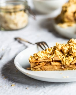 Ein Stück Apfelkuchen auf einem weißen Teller, der angeschnittene Kuchen aus der Springform in der Unschärfe dahinter