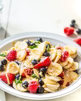 Pancake Cereals – Mini-Pfannkuchen in einer Frühstücksschüssel mit roten Beeren, Mandelmus und Minze, fotografiert seitlich auf hellem Untergrund.