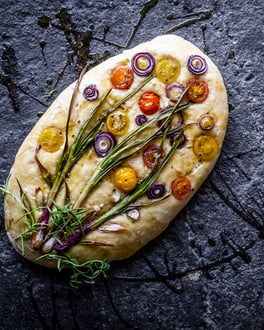 Focaccia Garden mit Tomaten, Rosmarin und Lauchzwiebeln drapiert wie eine Blume auf dem Grundteig auf einem Schiefer-Untergrund liegend.