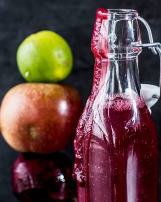 Glasflasche mit rotem Smoothie neben gestapeltem Turm aus ganzer Rote-Bete-Knolle, Apfel und Limette