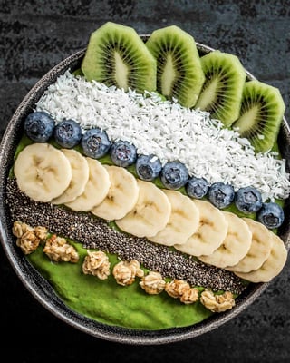 Grüne Smoothie-Bowl mit Topping-Streifen aus Kiwi, Kokosraspel, Heidelbeeren, Banane, Chia und Granola auf dunklem Untergrund