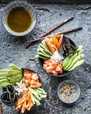 Zwei Sushibowls mit Karotten- und Gurkensticks, Avocadoscheiben, Reis, Lachsstücken und schwarzen Algen neben Essstäbchen auf grauem Stein