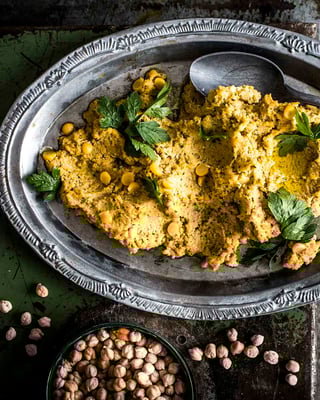 Einfacher Hummus, angerichtet auf silbernem, verzierten Teller, mit Löffel auf dem ovalen Teller, fotografiert auf dunklem, rustikalen Untergrund von oben. Kichererbsen sind zu sehen.