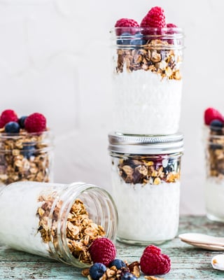 Granola mit griechischem Joghurt und Früchten in Schraubgläsern, neben Holzlöffelchen.