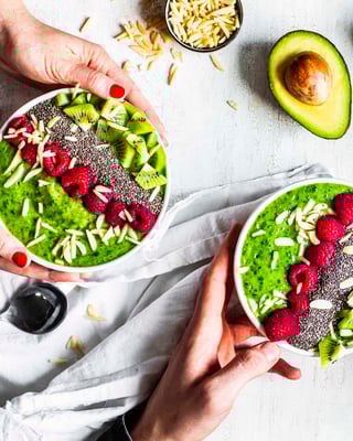 Zwei Hände, die jeweils eine Bowl mit grünem Smoothie, Himbeeren, Chiasamen, Kiwistücken und Mandelsplittern halten auf hellem Untergrund
