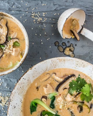 Asiatische Suppe mit Kokos, Shiitage und Huhn in Suppenschale, nebenliegend eine Suppenkelle auf grauem Untergrund.