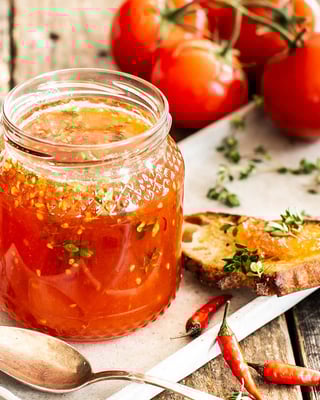 Roter Aufstrich im Glas mit Thymian, daneben ein Scheibe Brot mit dem Aufstrich bestrichen, Chilis und roten Tomaten als Deko.