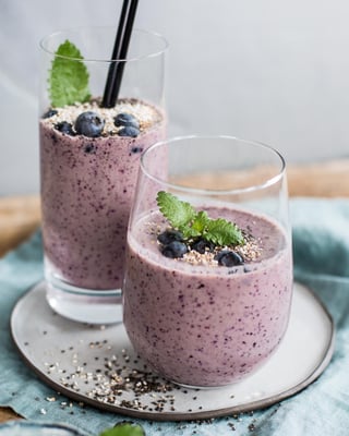 Zwei Gläser mit lila Heidelbeer-Smoothie getoppt mit Amaranth, Heidelmeeren und Minze auf türkisem Tuch