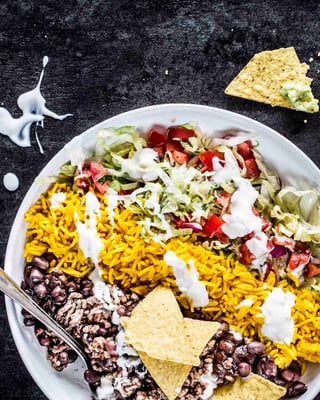 Burrito Bowl mit schwarzen Bohnen, Reis, Salat und Tomaten in Streifen angerichtet neben Guacamole und Joghurt in Schalen auf schwarzem Untergrund