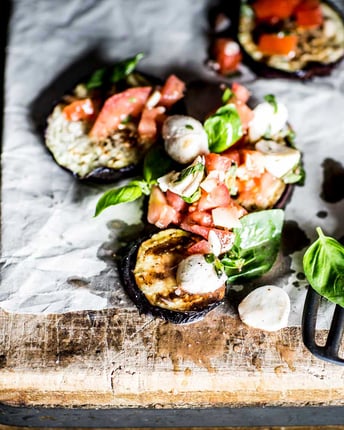 Gefüllte Aubergine mit Mozzarella, Tomaten und Basilikum auf Bckpapier und einem Holzbrettchen.