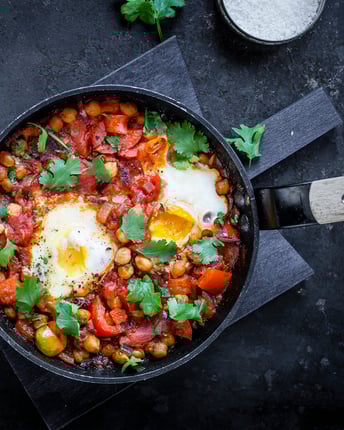 Die rote Mischung von Tomaten, Paprika, Chili und Kichererbsen wird mit zwei pochierten Eiern und Korianderblättern als Farbklecks aufgebrochen. Die bunte Pfanne steht auf dunkelgrauem Untergrund.