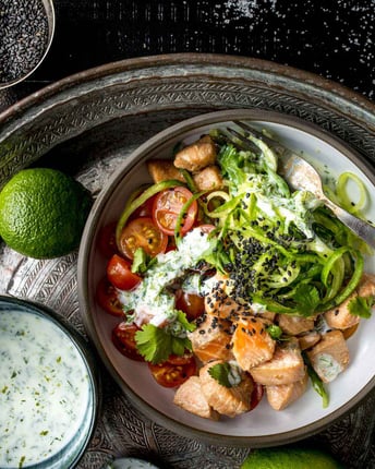 Schale mit Lachsstücken, Tomatenhälften und Gurkenspaghetti neben Joghurt-Dip und Limetten auf silbernem Tablett