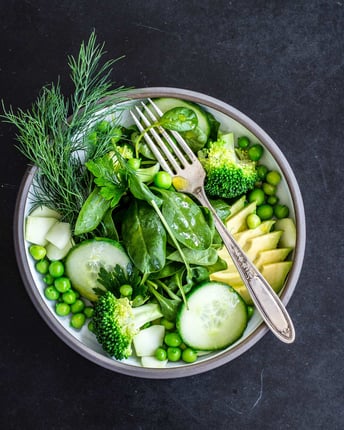 Buddha Bowl mit Spinatblättern, Gurkenscheiben, Avocado, Erbsen, Brokkoli und Dill mit Gabel auf schwarzem Untergrund