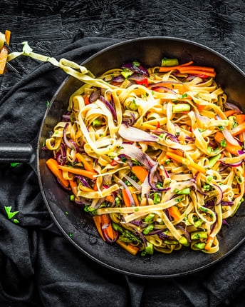 Farbenfroh wirkt dieses Pastagericht durch den grünen Spargel und die orangenen Möhrenstifte, die mit den Tagliatelle in der dunklen Schale vermischt sind. Dabei liegen Stäbchen, auf die ein paar Nudeln aufgerollt sind.