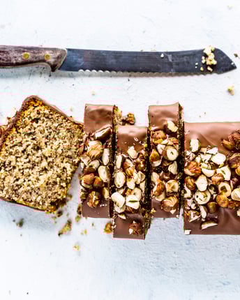 Nusskuchen mit Schoko-Glasur in Kastenform, von oben fotografiert, mit Haselnüssen dekoriert, angeschnitten auf weißem Untergrund. Brotmesser liegt daneben.