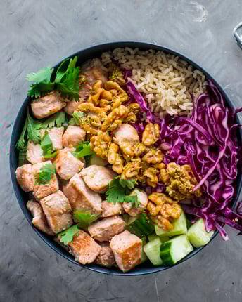 Bowl gefüllt mit Reis, Lachstücken, Rotkohl, Gurke und Walnüssen auf grauem Grund neben Walnusskernen