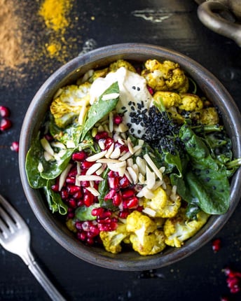 Curry-Zimt-Blumenkohl, Spinat, Granatapfelkerne und Mandelsplitter in einer Schüssel auf einem dunklen Untergrund auf dem Currypulver verteilt liegt.