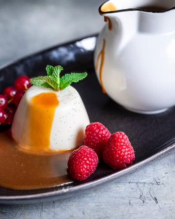 Klassische Panna Cotta mit Karamellsauce und Beeren auf schwarzem Teller auf grauem Untergrund
