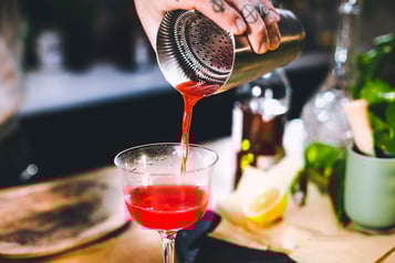 Eine Hand füllt den Jack Rose Cocktail mit einem Strainer aus dem Shaker in ein Cocktailglas.