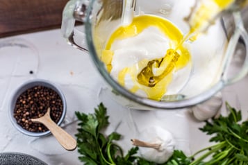 Standmixer auf mosaikweißem Untergrund mit Joghurt und Olivenöl und angestellten Chiliflocken
