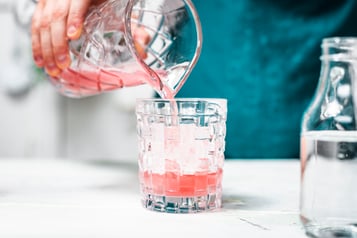 Der pinke Drink wird in ein Glas mit Eiswürfeln gegossen