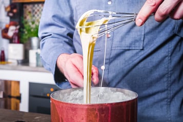 Cremige Käsesauce, die vom Schneebesen in den Kupfertopf fließt