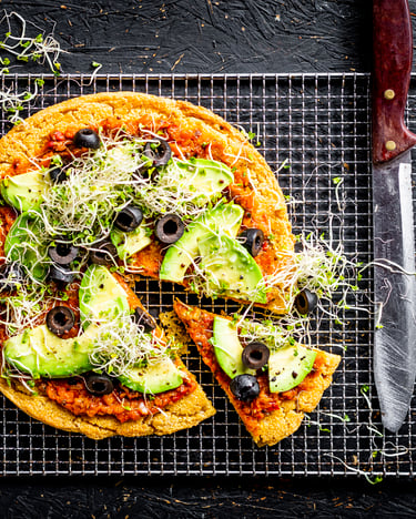 Socca Pizza mit Avocado und Oliven, mit Tomatengrund. Ein Achtel ist angeschnitten, Pizza wird von oben fotografiert und liegt auf einem Gitter.