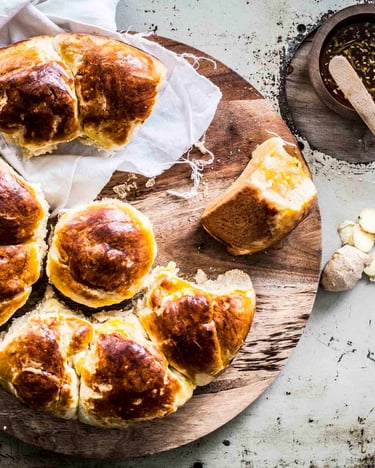 Luftige selbst gemachte zerteilte Brioche auf einem runden Holzbrett mit Ingwer-Honig in einem Schälchen auf grauem Untergrund.