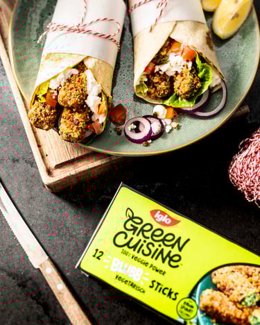Auf einem hellgrünen Teller liegen zwei gerollte Wraps, die mit buntem Gemüse und Spinat-Sticks gefüllt sind. Unten rechts in der Ecke liegt die grüne Verpackung der iglo-BLUBB-Sticks.