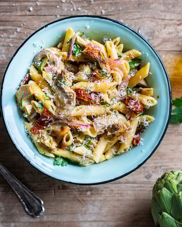 Penne mit Artischocken und Weißwein auf blauem Teller vor hellem Holzuntergrund