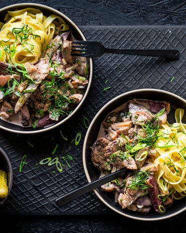Boeuf Stroganoff mit Tagliatelle angerichtet, auf zwei dunklen Teller, von oben fotografiert, daneben eine kleine Schale mit Gewürzgurken. Frühlingszwiebelringe liegen etwas verstreut auf dem dunkeln Untergrund.