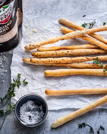 Grissini-Stangen auf weißem Backpapier mit Salz, Thymian, Olivenöl und Parmesan auf hellgrauem Betonuntergrund