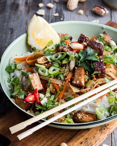 Phad Thai in einer Schale mit Gemüse und Tofu sowie Essstäbchen auf einem Holzbrettchen serviert
