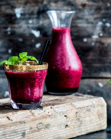Zwei Gläser mit Goldrand und Minze und Glaskaraffe mit pinkem Beeren-Smoothie vor dunklem Holzgrund