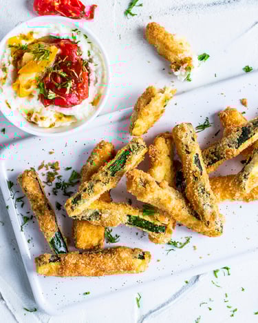 Zucchini-Pommes auf einem weißen Brett mit Feta-Dip und geschmolzenen Tomaten  in einer Schüssel daneben.