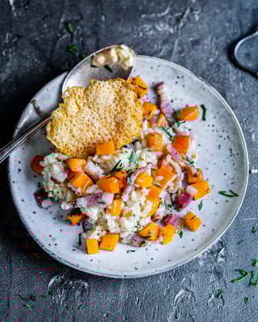 Kürbisrisotto mit Kürbiswürfeln, Guanciale und Parmesanchip auf rundem Teller und dunklem Untergrund, daneben ein Stück Parmesan und eine Reibe