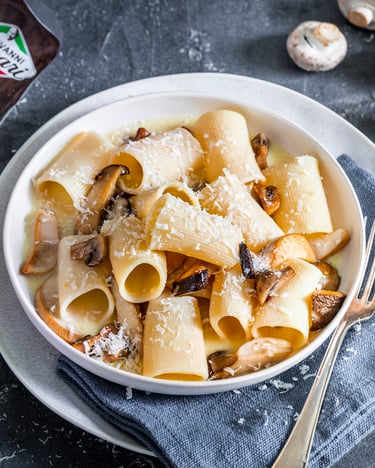 Paccheri mit Parmesansauce und Pilzen in weißem Teller auf dunklem Untergrund, im Hintergrund Parmesan und Champignons