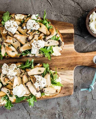 Zwei belegte Pilz-Stullen mit Salat auf einem Holzbrettchen auf grauem Untergrund.