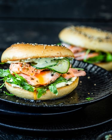 Aufgeschnittener und belegter Bagel mit Gurke, Rucola und pulled Lachs neben einem Glas Ahornsirup