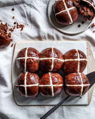 Schokoladige Hot Cross Buns mit hellem Kreuz auf einem weißen Tischtuch.
