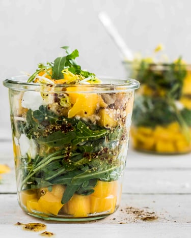 Mango-Salat mit Rucola im Glas mit Besteck auf weißem Holzuntergrund.