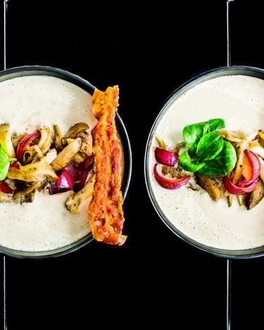 Zwei Schüsseln mit Pilzsuppe getoppt mit Speck, Zwiebeln und Feldsalat auf schwarzem Untergrund