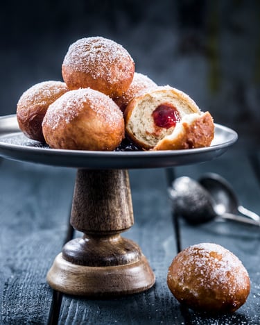 Mini-Berliner mit Erdbeerkonfitüre auf einem Serviertablett vor dunklem Hintergrund. Im Hintergrund liegt in der Unschärfe ein offenes Teesieb.