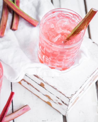 Zwei Gläser sind mit dem pastellig-pinken Drink gefüllt und mit jeweils einer Stange frischem Rhabarber dekoriert.