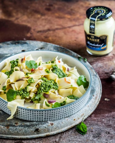 Pasta mit Spinat-Pesto und Chili angerichtet in einer Schüssel mit Karo-Muster auf einem metallenen Unterteller mit Besteck daneben. Ein Senfglas und ein Glas mit Pesto stehen ebenfalls im Bild.