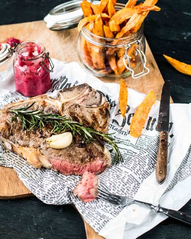 Angeschnittenes T-Bone-Steak auf Zeitungspapier mit Süßkartoffelpommes und Beeren-Ketchup im Glas.