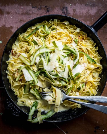 Spargel-Safran-Pasta in dunkler Pfanne, mit Spargelstreifen, fotografiert auf braunem Holz, Besteck liegt in der Pasta, ebenfalls zu sehen ist eine Reibe mit Parmesan.