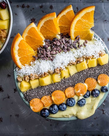 Mango-Smoothie-Bowl mit Toppings in Streifen aus Orangenspalten, Kakaonibs, Kokosraspel, Mango, Chiasamen, Physalis und Heidelbeeren auf dunklem Grund
