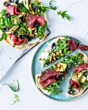 Drei kleine Pizzen, die üppig mit Ruccola, Schinken und Zucchini belegt sind, sind auf einem Gitter und einem blauen Teller serviert.