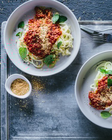Kohlrabi-Spaghetti mit Graupen-Bolognese, angerichtet mit gehackten Mandeln in Pastatellern auf einem silbernen Tablett.